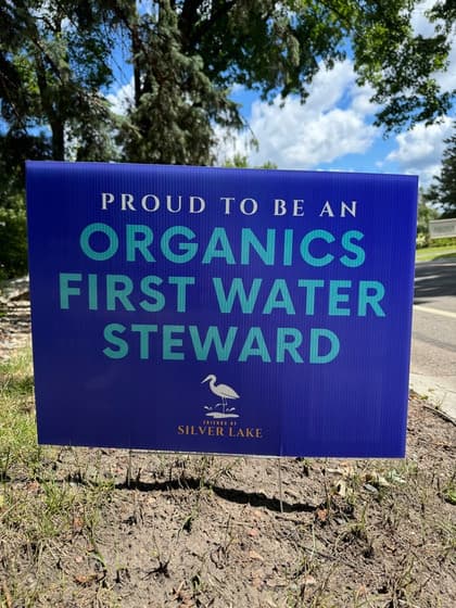 A blue yard sign reading “Proud to be an Organics First Water Steward”